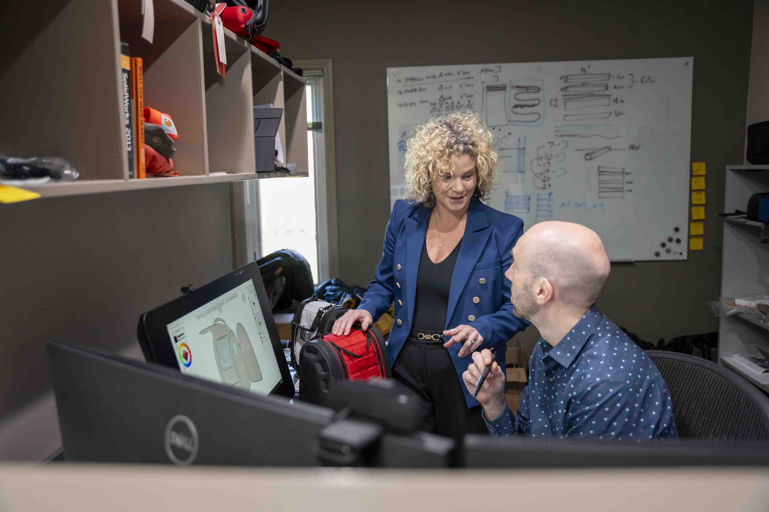 woman holding prototype of backpack discussing with man in front of two monitors