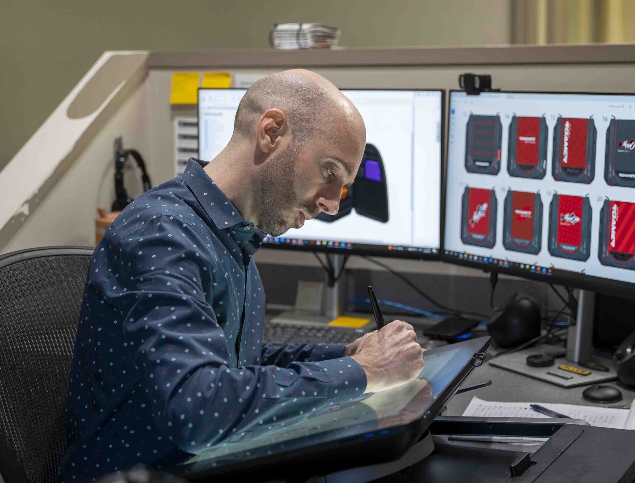 man sketching on a device with multiple monitors behind him showing backpack drawings